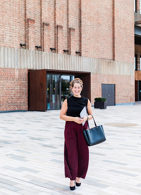 Zoe Thompson - white female, brown hair, red trousers, black top walking outside Battersea Station London with bag and book