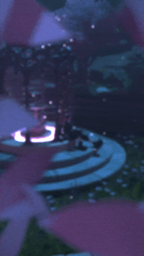 A person sits on stone steps in a garden-like setting, dressed in a black outfit with large boots and wearing headphones. The background features a classical structure with columns and a canopy, adorned with vines and surrounded by lush greenery. Purple-lit trees cast a soft glow, and ambient lighting creates a serene, mystical atmosphere.