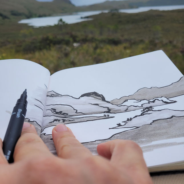 Artist's hand holding a pen next to  the sketch in foreground, with blurry landscape in the background