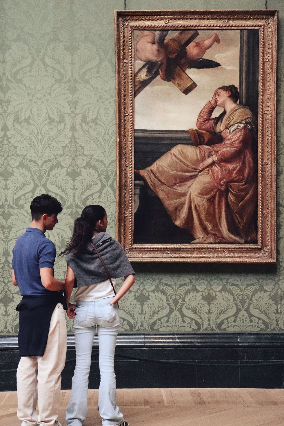 Two people stand closely together, admiring a large framed painting in a museum gallery with ornate wallpaper. The artwork features a reclining woman and an angel or cherub holding a cross, bathed in soft, classical light.