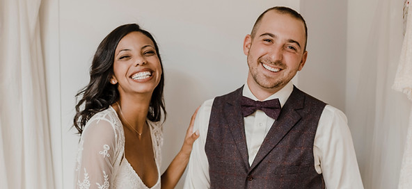 A soon to be married couple stands together in their wedding outfits looking at the camera and smiling with a laugh.
