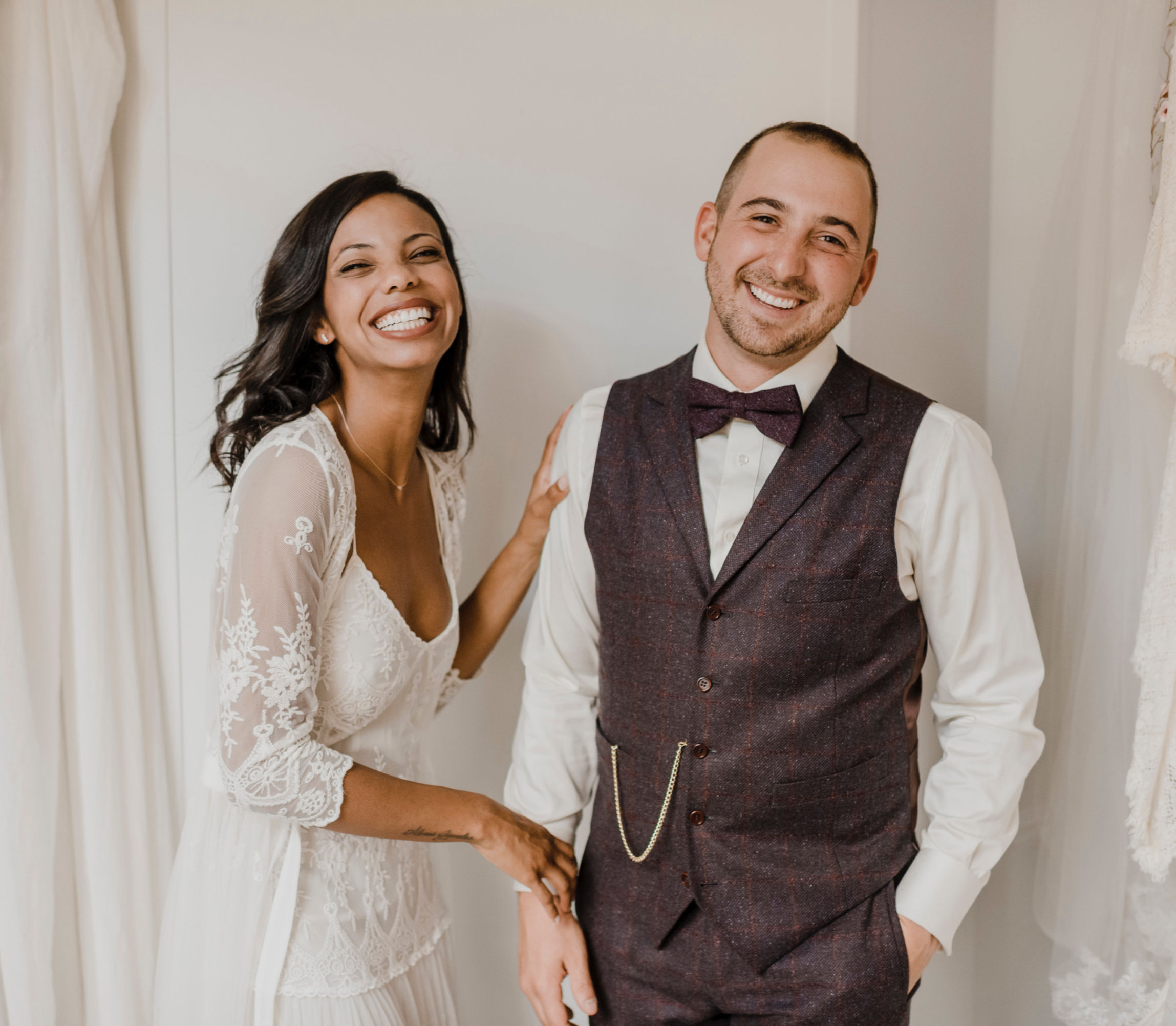 A couple stand against a white background laughing during their wedding photoshoot.