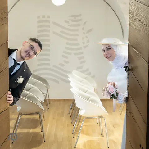 Ein Brautpaar schaut lächelnd durch eine halb geöffnete Holztür in einen hellen Raum. Die Braut trägt ein weißes Kleid mit Hijab und Hut und hält einen kleinen Strauß rosa Rosen. Der Bräutigam trägt einen dunklen Anzug mit Krawatte und Boutonniere. Im Hintergrund stehen mehrere weiße Stühle in Reihen auf einem Holzfußboden, an der Wand ist eine stilisierte Figur als Dekoration zu sehen.