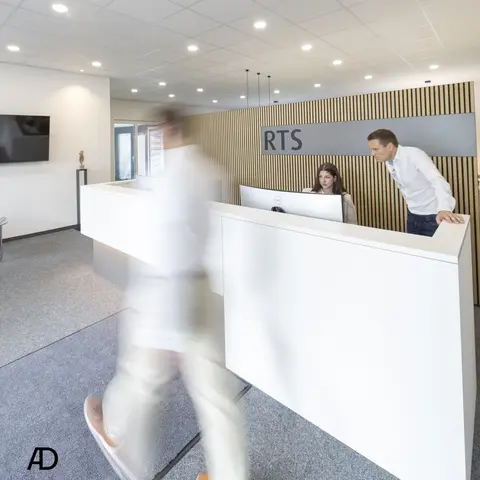 Heller Empfangsbereich eines Büros mit modernem weißen Tresen. Eine Frau sitzt am Computer, ein Mann lehnt sich daneben und schaut auf den Bildschirm. Im Vordergrund geht eine Person unscharf durchs Bild. An der Wand steht das Logo „RTS“ vor einer Holzwand mit vertikalen Streben.