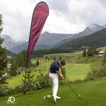 Ein Golfer in weißer Hose und dunklem Shirt schlägt auf einem Berggolfplatz ab. Neben ihm steht ein rotes Banner mit der Aufschrift ‚Burg Golf Trophy‘, im Hintergrund grüne Hügel, Wälder und Berge unter bewölktem Himmel.