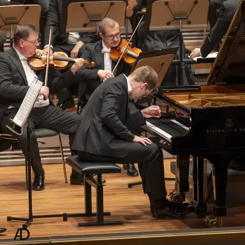 Pianist in schwarzem Anzug spielt intensiv auf einem Steinway-Flügel während eines Konzerts, begleitet von Streichern des Orchesters im Hintergrund.