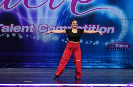 Teen dancer in black top and red pants performs tap dance on stage at dance competition.