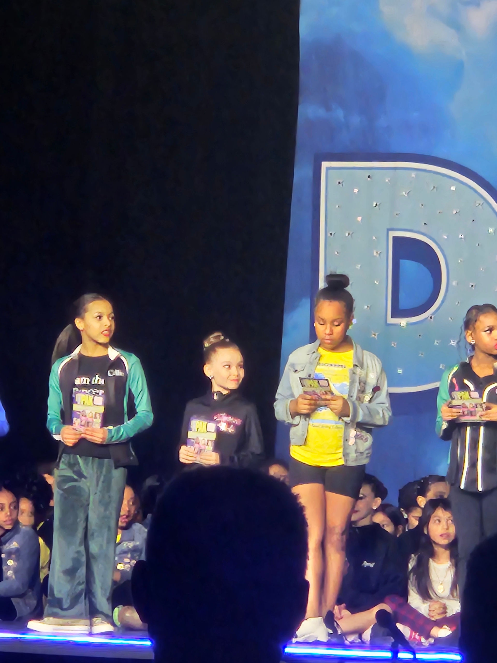 Young dancers holding awards on a stage with the letter D in the background.