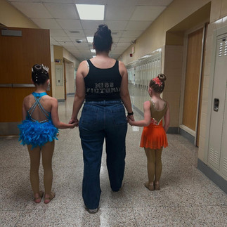Woman with two young dancers walk down hallway, wearing costumes.