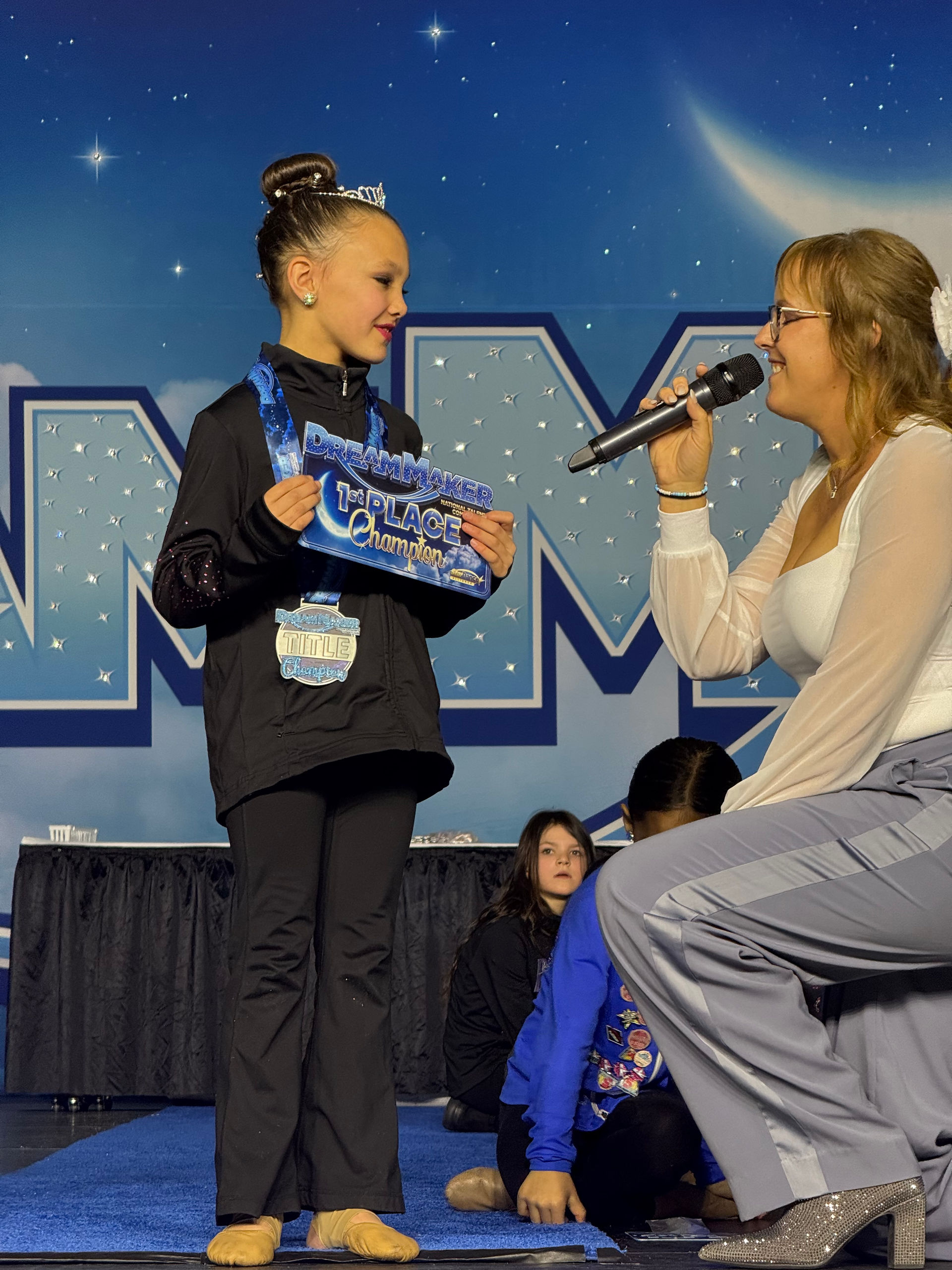 Young dancer receives award on stage with a woman and microphone.