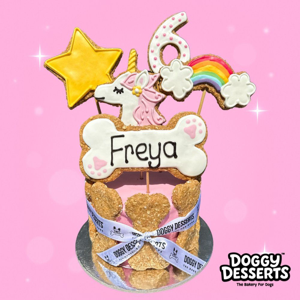 Pink dog birthday cake topped with hand-iced unicorn, rainbow, star, and number biscuits, surrounded by bone biscuits.