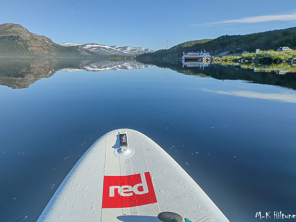 Paddleboard with "Red" logo on calm Kilpisjärvi lake, surrounded by mountains, boat docked on right. Clear blue sky and serene mood.