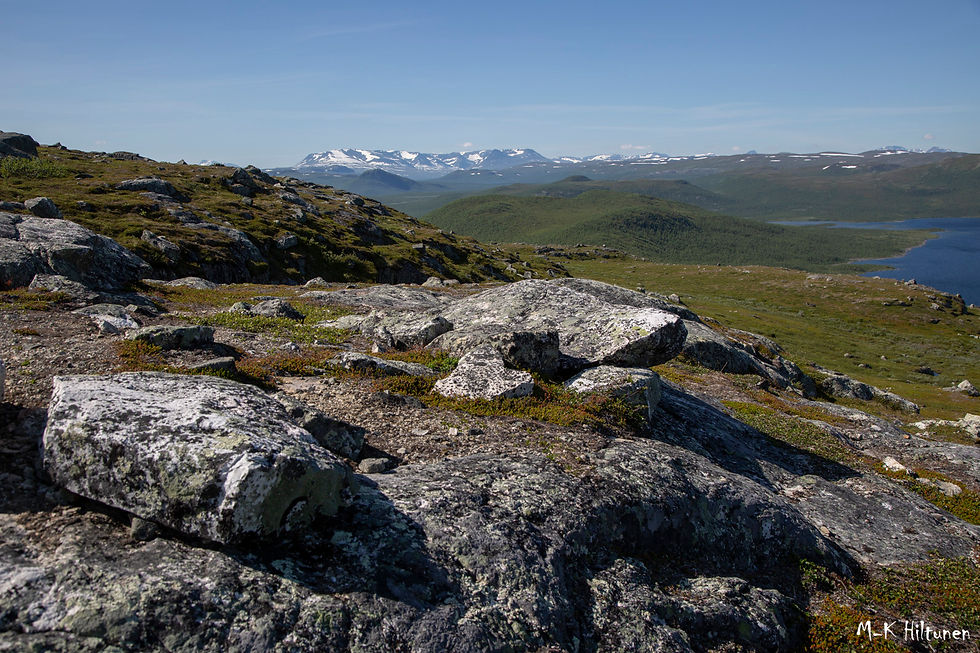 Rocky landscape with green hills, distant snow-capped mountains in Norway, and blue sky. Lake Kilpisjärvi visible to the right. Calm, serene setting.