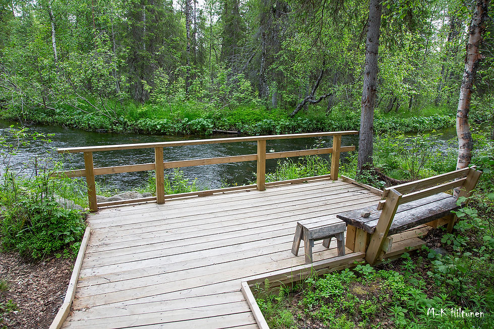 Wooden deck with bench and stool by a flowing river Orajoki in a lush forest. Green foliage surrounds the peaceful setting; initials in corner.
