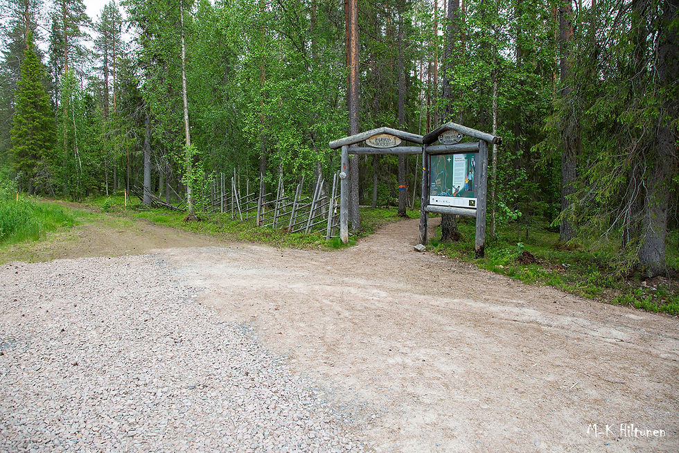 Forest path entrance with wooden archway and sign of "Paavon polku", bordered by a rustic fence. Lush green trees in the background, gravel path foreground.