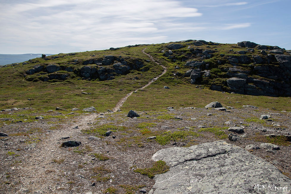 Rocky Leutsuvaara hill with a narrow dirt path winding up. Grass and scattered rocks under a cloudy blue sky. Calm and open landscape.