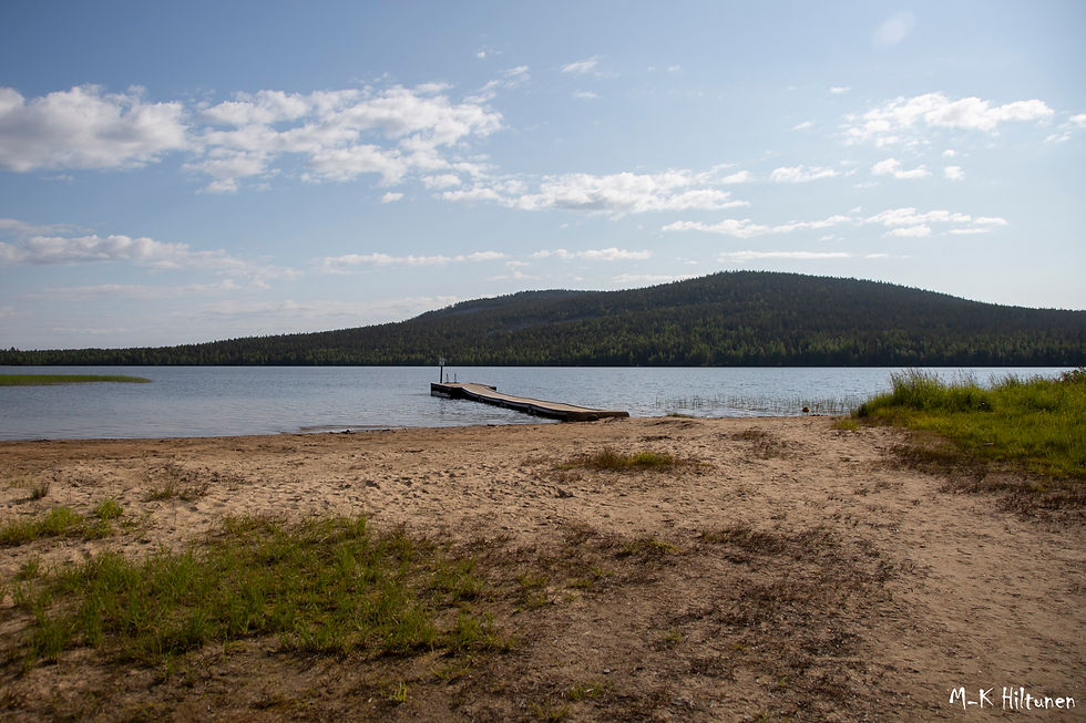 Tranquil lake with a wooden dock extending into the water. Sandy beach in the foreground, forested Soutaja-tunturi in the background under a clear sky.