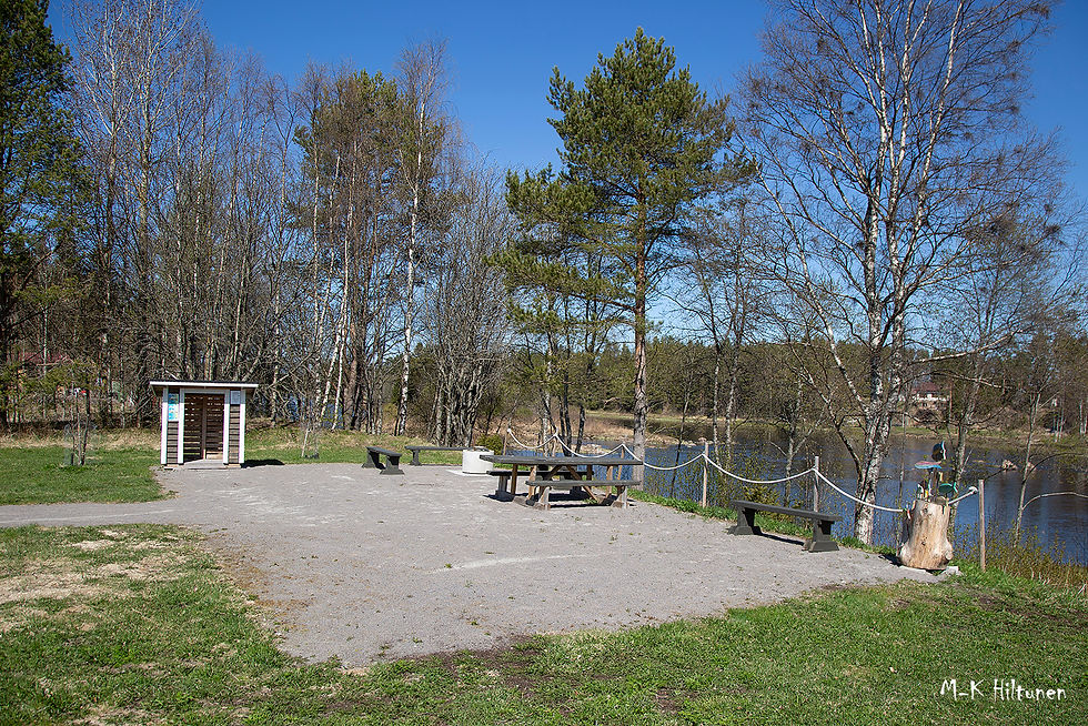 A park in Siikajoki with picnic tables and benches beside a pond, surrounded by trees. A wooden structure is on the left. The scene is calm and sunny.