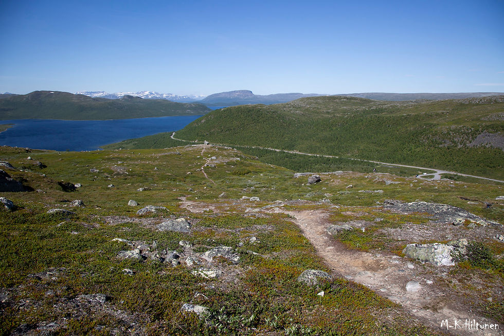 Mountain landscape from Leutsuvaara with a winding dirt path, green hills, and a deep blue lake under a clear blue sky. M-K Hiltunen signature in corner.