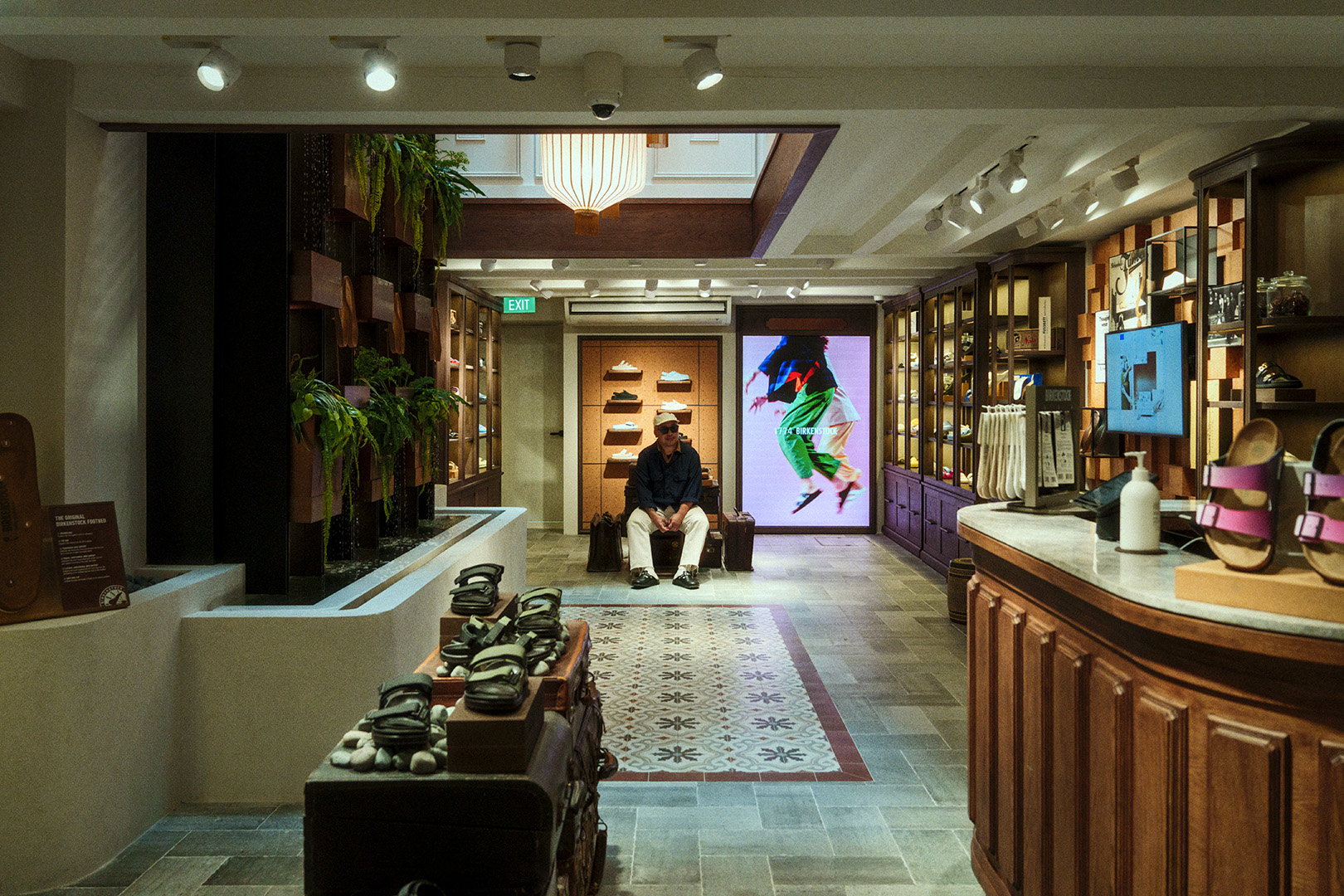 man sits on bench facing camera in distance surrounded by display items inside the House of Birkenstcok at duxton road with wood-rich and elegant interior decor in mix of modern and traditional asian style