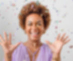 Woman in a purple top joyfully throws colorful confetti. She smiles widely, hands raised, against a plain white background.
