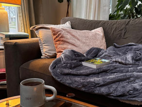 Cozy slow living reading nook with a lit candle, warm tea, soft blankets, and a book on the couch, creating a peaceful ritual of rest at home.
