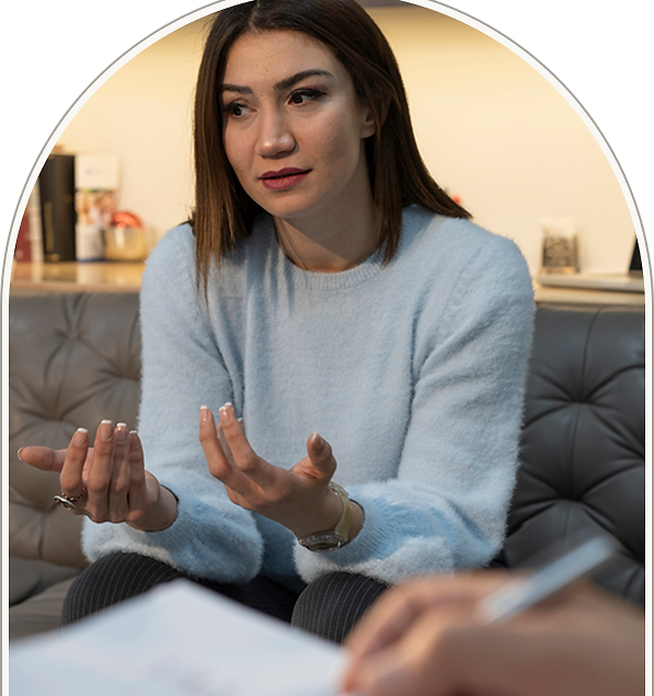 Woman in a light blue sweater talking during a counselling session, expressing emotions while seated on a sofa in a therapy room.