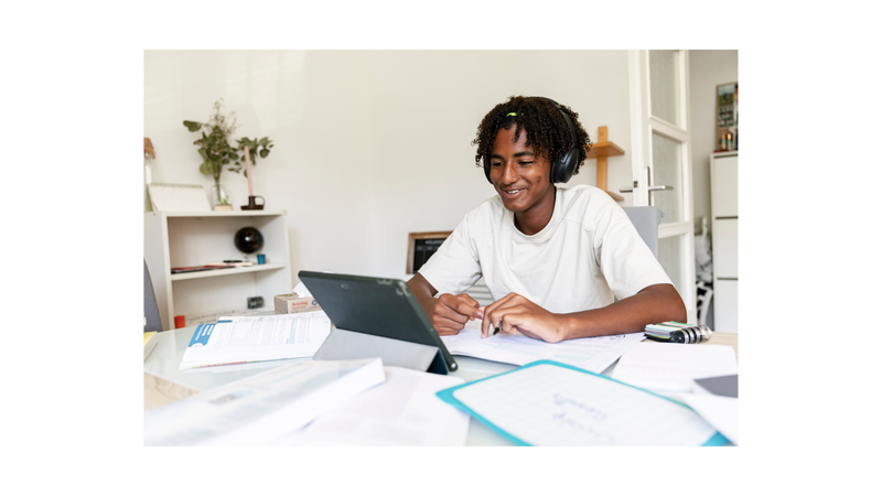 A high school male at home sitting in front of his tablet with school papers surrounding him.