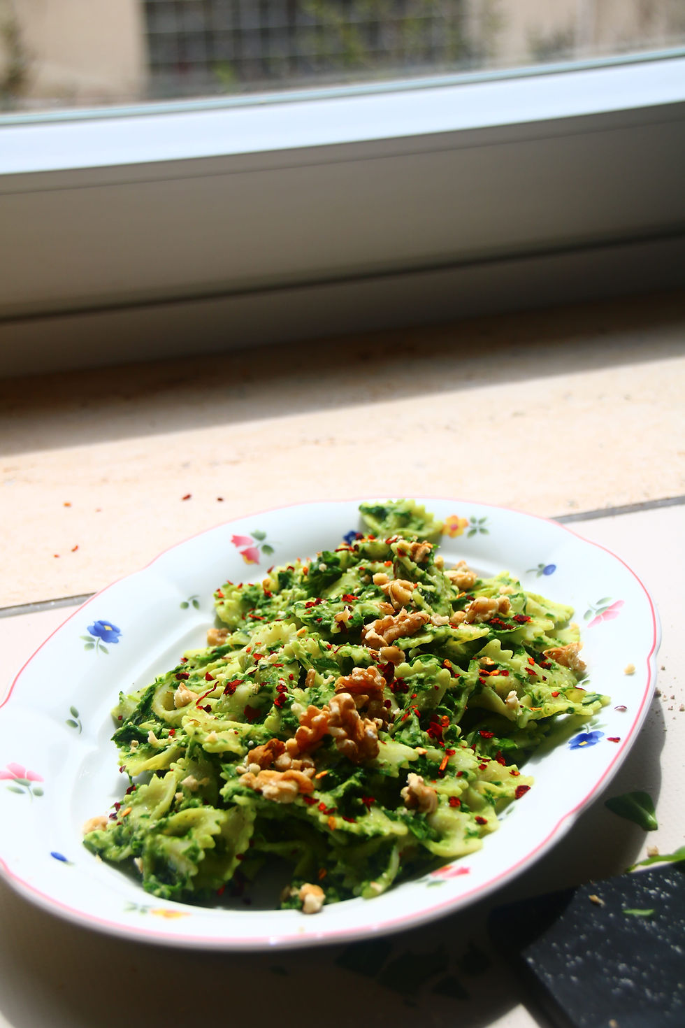 Plate of green pasta with walnuts and red pepper flakes on a floral dish by a sunlit window.