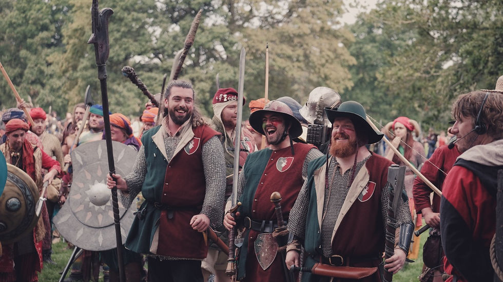 People in medieval costumes with shields and spears, smiling during an outdoor event. Trees in the background, lively atmosphere.