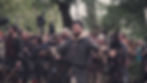 Man in chainmail stands with arms outstretched in a forest, surrounded by people in medieval costumes. Mood is serious, setting is lively.