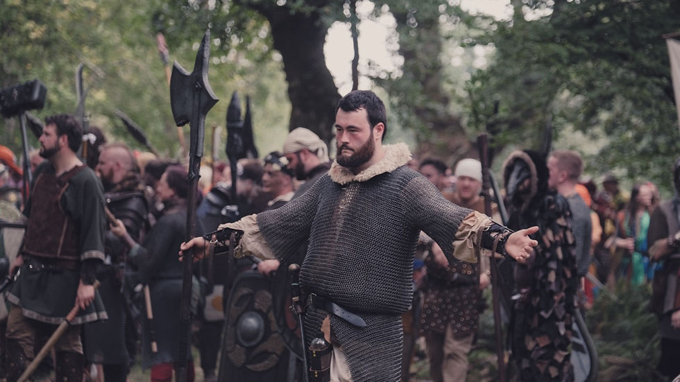 Man in chainmail stands with arms outstretched in a forest, surrounded by people in medieval costumes. Mood is serious, setting is lively.