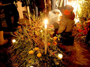 A child in warm clothes sits by a grave adorned with marigolds and candles, among standing people, in a dimly lit cemetery on the Day of the Dead.