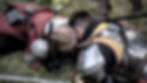 Two armored individuals crouch on grass, leaning close together. One wears red, the other yellow. The scene is intense and somber.