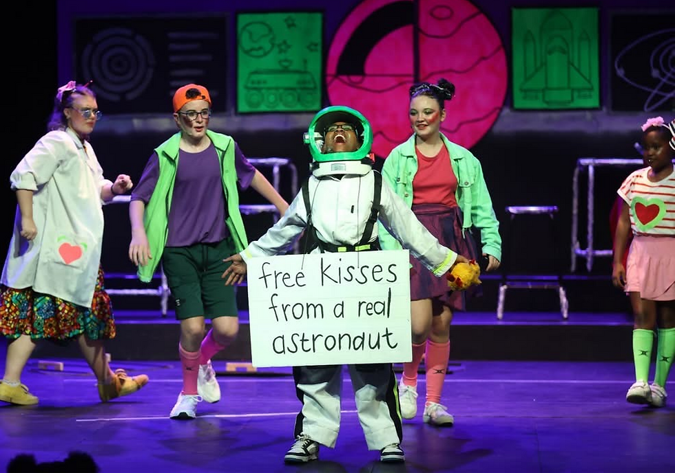Kids on stage dance around a child in an astronaut costume holding a sign reading free kisses from a real astronaut