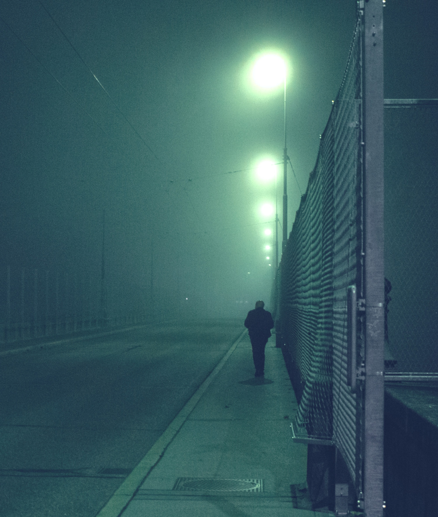 Lone person walks along a foggy, empty road beside a chain-link fence under glowing streetlights at night.