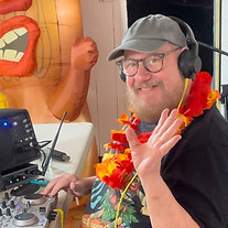 Male DJ waving and smiling. He has a hat and glasses on. At a luau party.