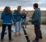 A group of people watching as a woman demonstrates how to use a pole to take water samples.