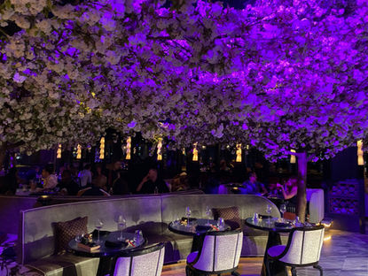 Restaurant with patrons under blooming tree, purple lighting. Elegant tables set with wine glasses. Cozy, vibrant atmosphere. Personal Escape Travel