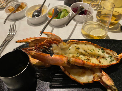 Wine and whiskey glasses on a table with cruise magazines. A plate holds a caramel pastry and chocolate dessert, set in a modern lounge.