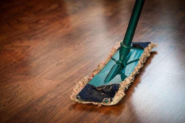 A green mop cleans a polished wooden floor. The setting is tidy, with a focus on the mop's worn blue and beige fabric.