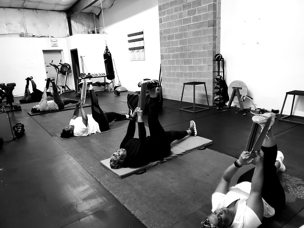 Five people in a gym lying on mats doing leg stretches with resistance bands. The setting is bright, with exercise equipment in the background.