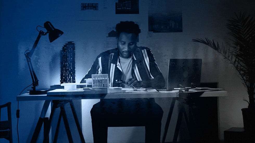 Man in a striped shirt studies architectural model at desk. Blue light ambiance, laptop open, modern lamp and plant in background. Text reads "Reignite Your Passion"