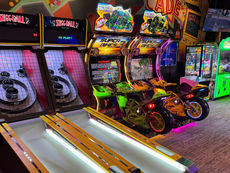 Arcade with skee-ball and motorcycle racing games, vibrant lights, and colorful machines. Neon signs and plush claw machines in view.