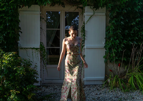 Woman in floral dress outside of white door with foliage background; Two Waves Creations