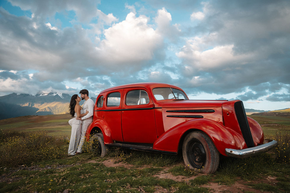 Andrew and Lexis embracing beside a vintage red car during their engagement photoshoot in Peru’s Sacred Valley, with the Andes Mountains and dramatic clouds in the background.