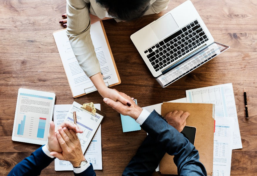 image of a woman shaking hands with a man with a laptop in front of her and a clipboard in front of another man.