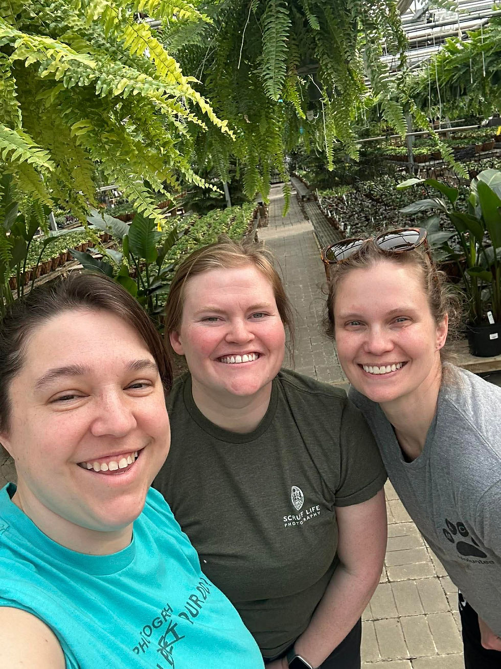Three smiling people taking a selfie in a lush greenhouse, surrounded by greenery. They wear casual shirts with text, showing a joyful mood.