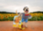 Dog dressed as artist with beret and bowtie, standing by easel with sunflower painting, surrounded by a vibrant field of sunflowers.