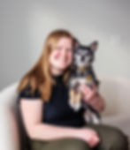 Woman smiling, holding a small black and white dog in a cozy room with a white wall. The dog's collar is gold. Calm and happy mood.